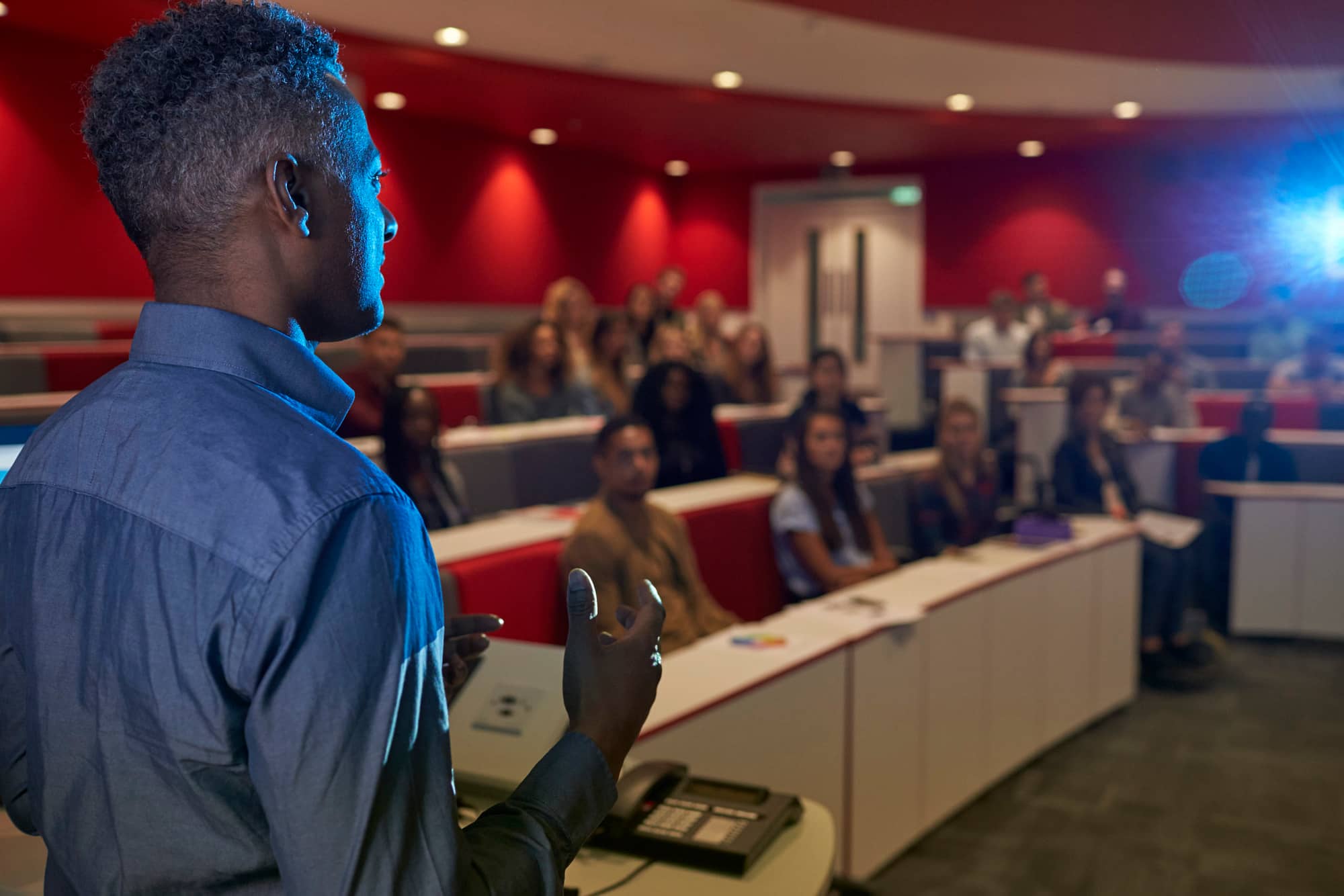 A male of darker complexion is presenting to a multi-level conference room full of audience members.